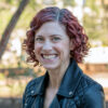 A woman with curly red hair and a big smile in a black jacket looks straight ahead with her body angled camera left.