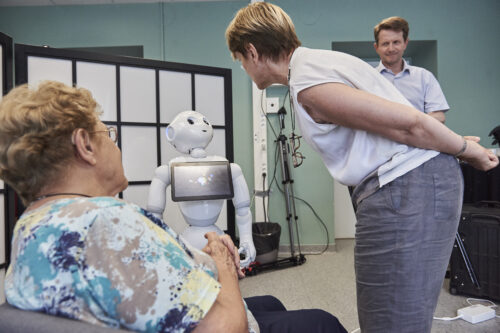 Three human researchers surround a white robot with a screen on its chest. The robot looks up to meet the gaze of one of the researchers.