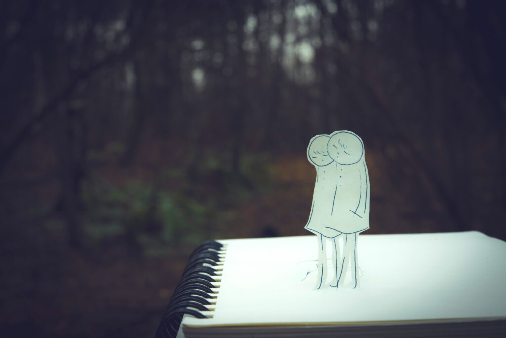 An image features a spiral notebook page against a blurry background of a forest. A paper cutout of a drawing of two people walking side by side stands on top of the paper.