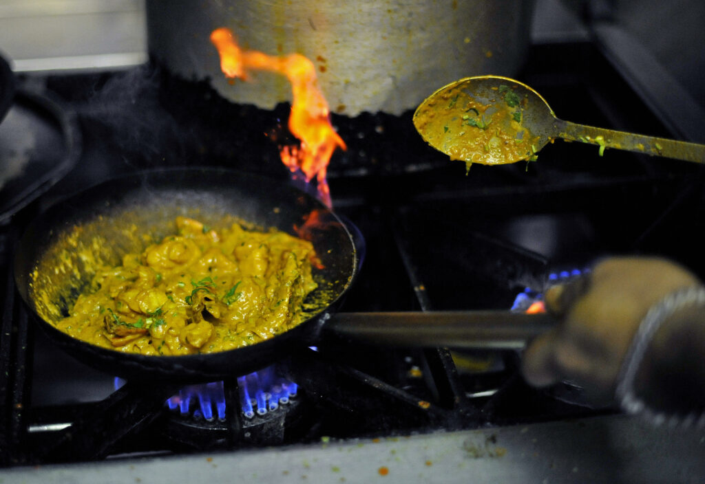 A person holds the handle of a frying pan filled with a yellow stew on a gas stovetop.