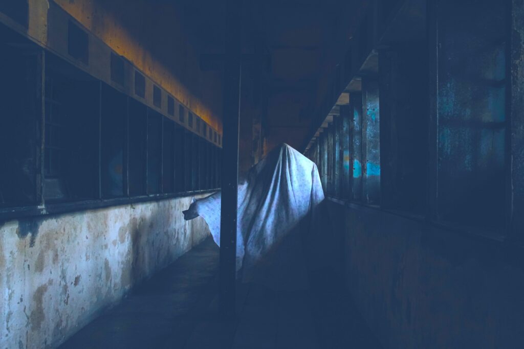 A human figure with a large white cloth draped over them stands in a long, dark corridor lined with windows on one side.