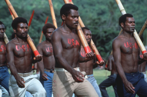 With forest in the background, shirtless people with "USA" painted in red on their chests wear a mix of jean and khaki bottoms. Each person holds a long bamboo stick in their left hand, balancing it on their left shoulder. The group steps forward in unison.