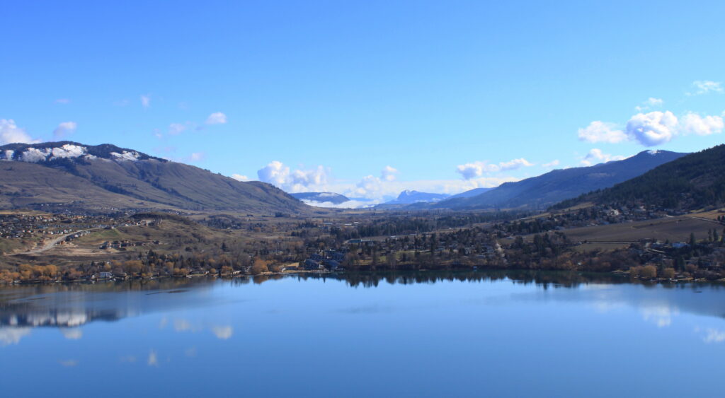 A photograph features a landscape with a large body of blue water in the foreground encircled by trees, small homes, and a few roads. The hills on which the latter sit ascend into mountains in the distance against blue skies.
