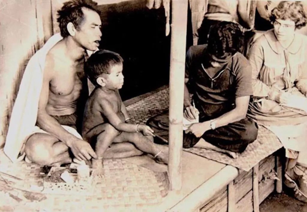 A black-and-white photo features a man and child sitting next to another man who sits cross-legged and takes notes. Behind him, a woman also takes notes.