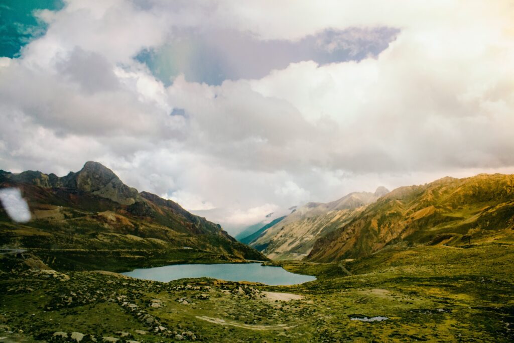 A photograph features a landscape lush with green grass with a body of water in the center and a large mountain range in the background. The mountain range is cut in half by a valley and topped with a sky full of white clouds.