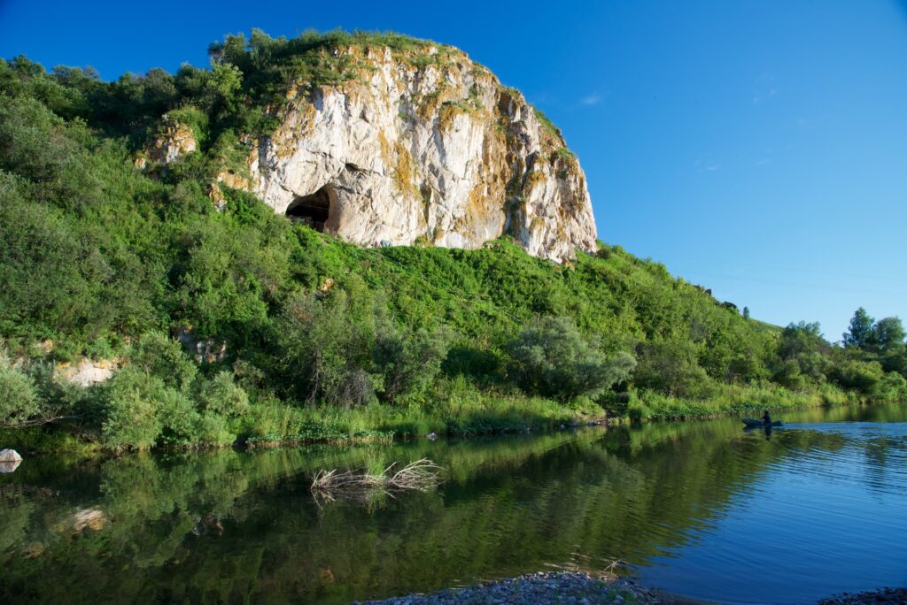 A body of water lies in front of a broad expanse of trees that surround a large rock buttress that holds a cave.