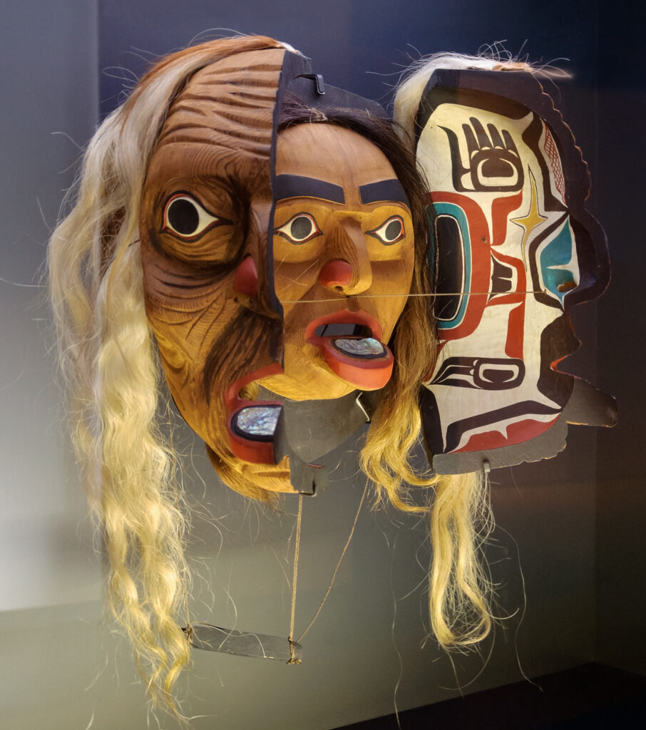 An elaborate mask is divided into three sections, depicting an older person with long gray hair on the left, a person with dark hair sticking out their tongue in the center, and a colorful abstract mask on the right.