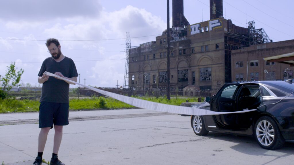 A person stands to the left side of a photograph, holding a long paper receipt that trails into a black car. A large building and green lawn sit in the background.