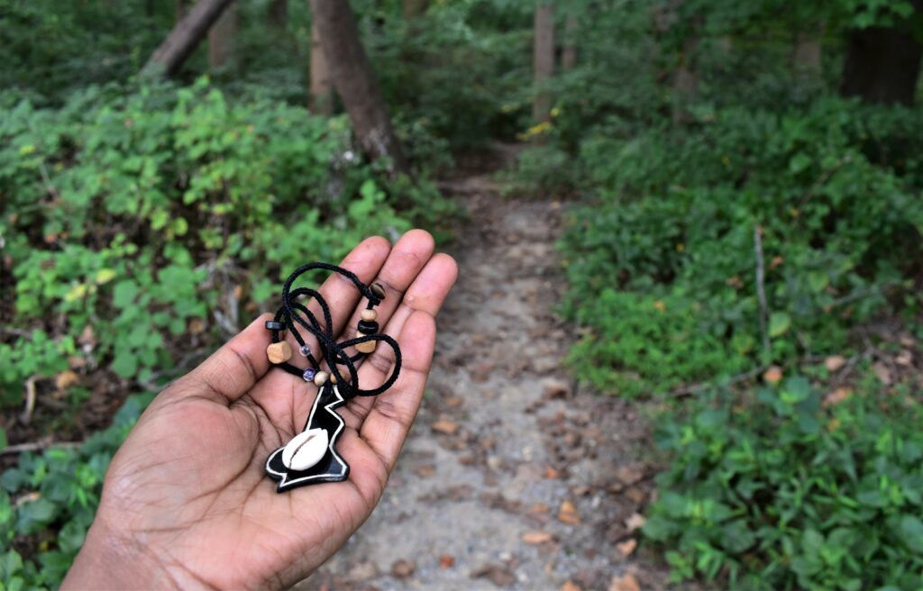 A photograph features a hand holding up a necklace with a black pendant with a white cowrie shell in its center. The shrub-lined entrance to a wooded area of tall trees fills the background.