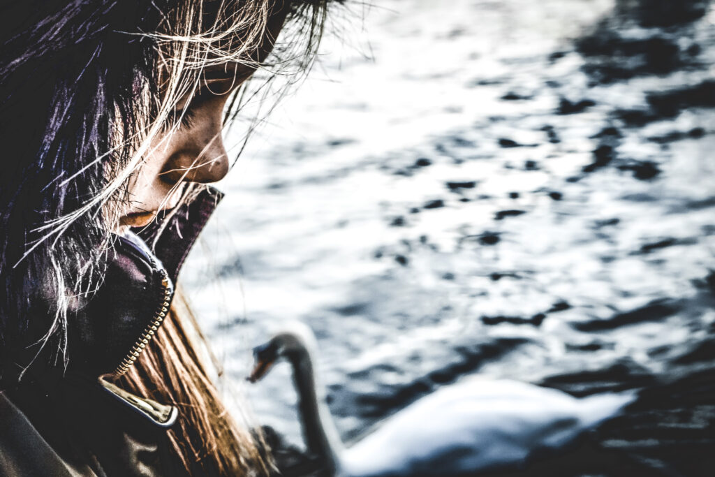 A close-up image features the profile of a child with long, stringy hair and a brown coat on the left side of the picture with a swan, sandy shoreline, and water in a blurred background.