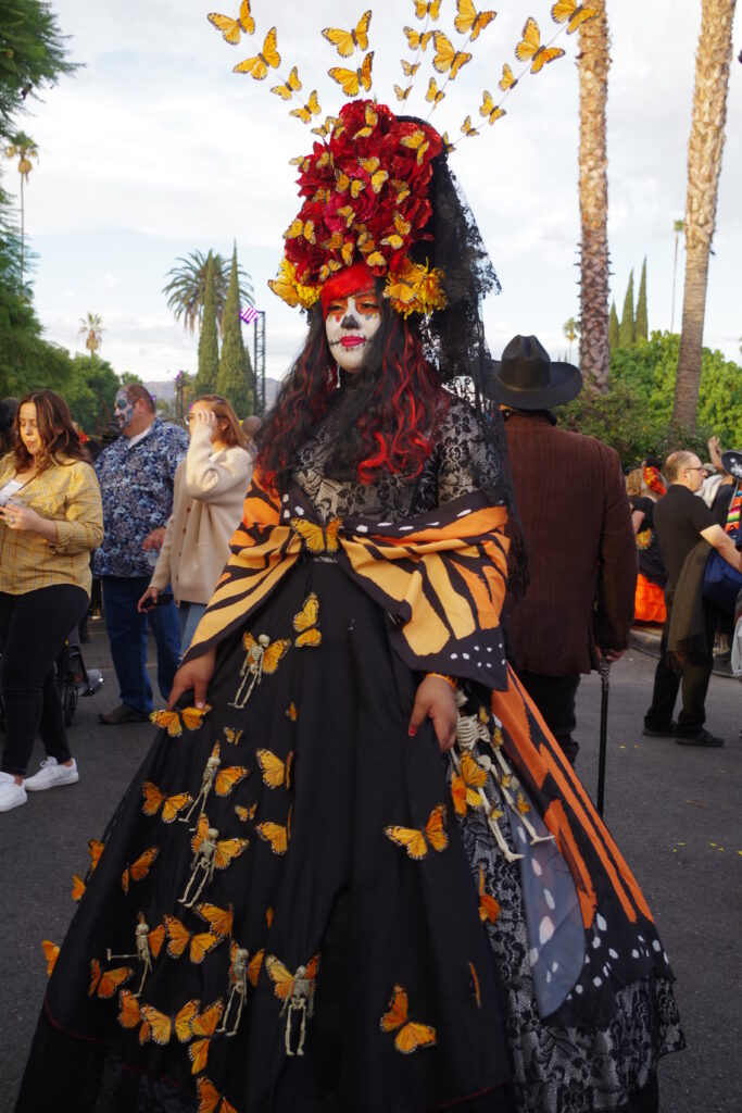 A photograph features a person posing with a dress with a full petticoat covered in orange and black butterflies. They also wear an orange and black shawl and a headpiece of orange and black butterflies, red roses, and black lace.