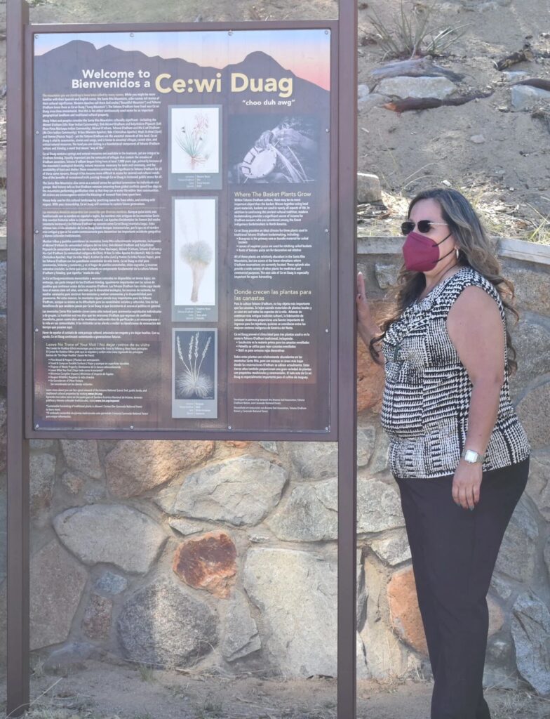 A woman stands next to a sign that reads “Welcome to / Bienvenidos a Ce:wi Duag, followed by lengthy text and images.