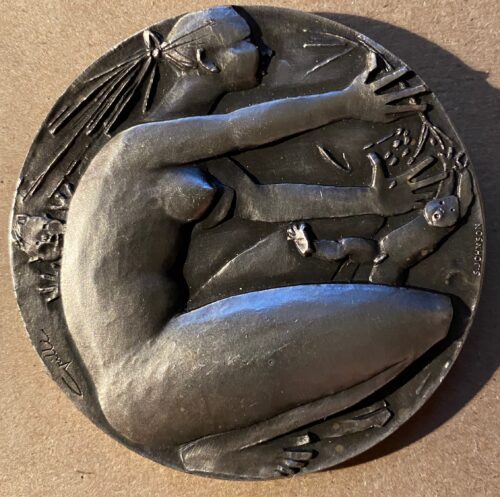A close-up image features a large, dark-gray silver medal embossed with the profile of a crouching naked woman reaching for a child, with another child appearing behind her.