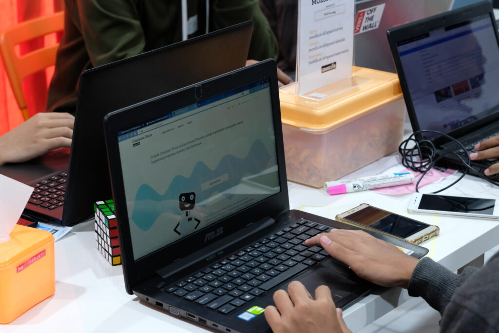A laptop screen features an automaton graphic and a wavy shape in the background. A person types on the laptop’s keys. Several people sit nearby, working on their own laptops.