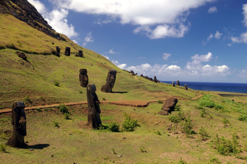 Head-shaped stone statues are scattered across a grassy hill with blue sky and ocean in the distance.