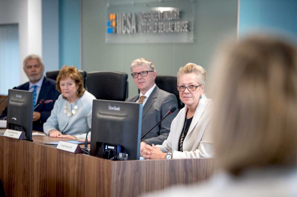 A photograph features four people in business attire seated at a wooden table that holds several notepads and two black computers. They look to their left at a person who appears blurry in the right corner of the foreground.