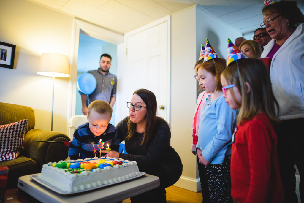 In a room with a lamp and sofa, a person props up a young child over a rectangular cake with burning candles stuck into it. The child blows at the candles while several other children and adults watch.