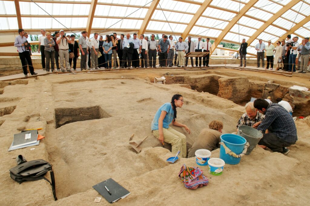 A few people in T-shirts and khakis sit on a dirt area with square holes dug into it. In the background, a group of people wearing business attire look on.