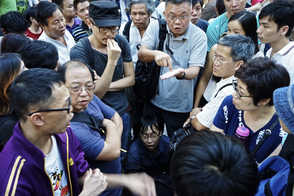 Several people stand in a circle in a public area, facing inward. Some point at other people and others lean forward while others cross their arms. One person even crouches under others to enter the scene.