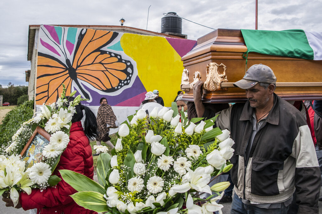 A photograph features a person holding a photograph brimmed with white flowers followed in procession by a larger white flower bouquet, and people carrying a wooden coffin with a flag on top. A large painted image of a monarch butterfly surrounded by other colors hangs in the background.