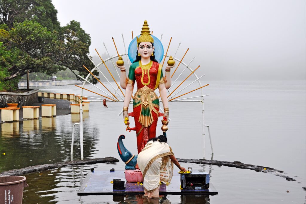 A photograph features a woman in a white sari bending down to place objects at the base of a statue. Erected on one side of a large body of water, the statue depicts a female deity with four arms wearing ornate red and green robes and holding multiple golden orbs.