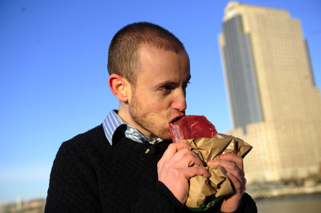 A photograph features a person in a striped, collared shirt and sweater as they bite into a piece of raw meat that they are holding in a paper bag.