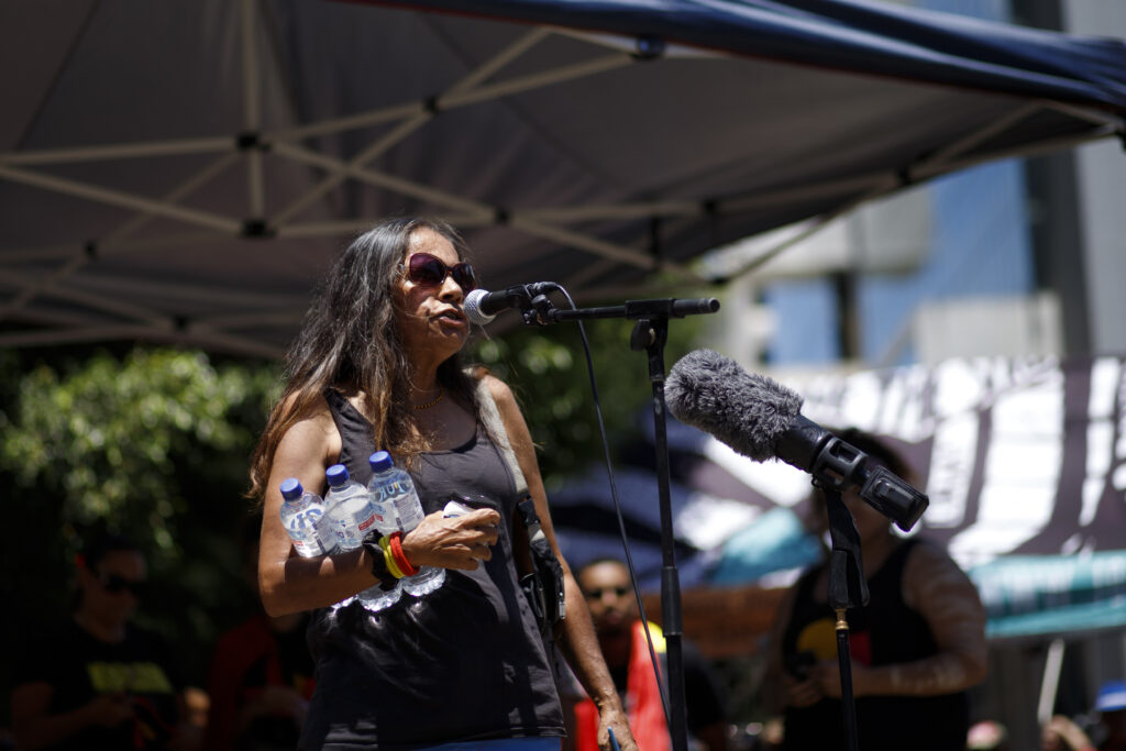 A woman in a black tank top stands at a microphone on an outdoor platform while holding three bottles of water.