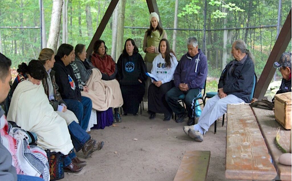On flattened earth under a wooden awning, a woman in a sweatshirt sits centered in an arc of other seated people.