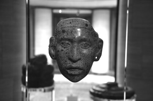 A black-and-white photograph features a stone carving that resembles a person’s face inside of a glass box display.