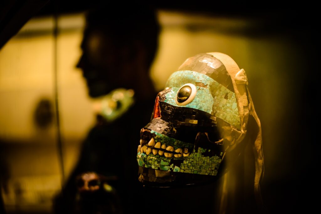 A mask that resembles a skull with stripes of teal across the eye and mandible sits behind glass that reflects the shadow of a person.