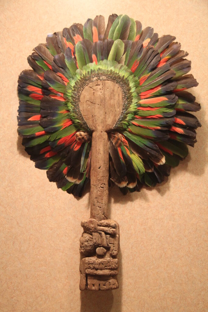 A fan with a handle made of carved wood topped with red, green, and black feathers hangs on a wall.