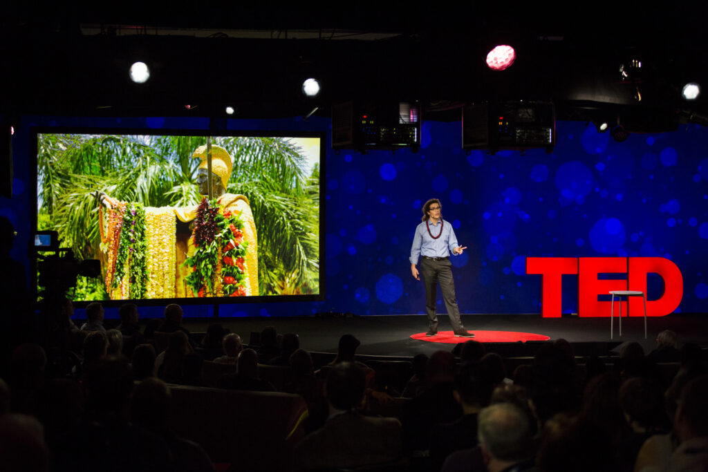 A person with long, curly hair wears glasses, a blue button-up shirt, and black slacks, and stands on a red carpet on stage. A screen behind him displays an image of a statue of a person in colorful robes and a headpiece with a series of leis, or garlands, on their arm.