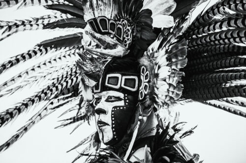 A black-and-white photograph features the head of a person with patterned face paint and a large headpiece adorned with feathers.