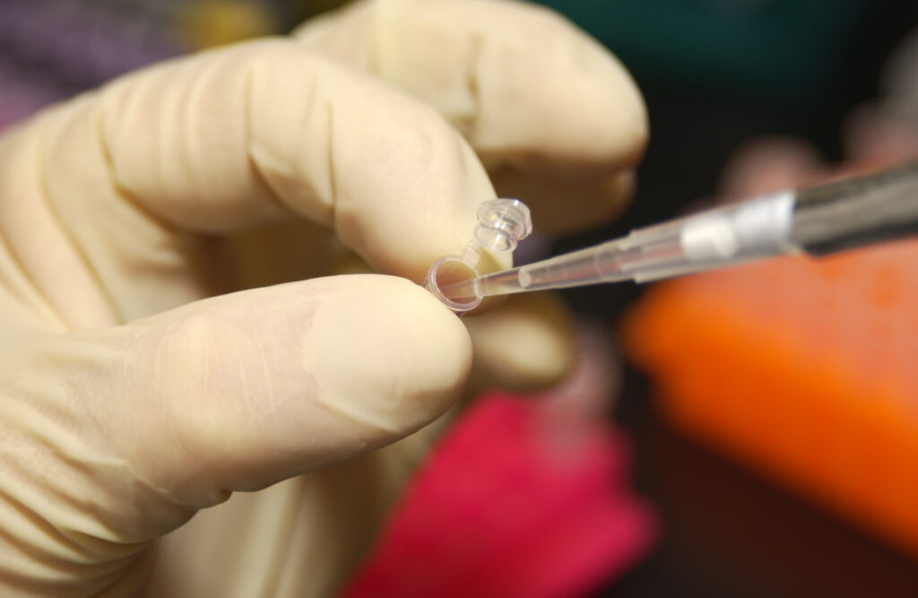 A close-up image features a hand covered with a white latex glove holding a tiny test tube as it catches drops of small clear liquid from a pipette.