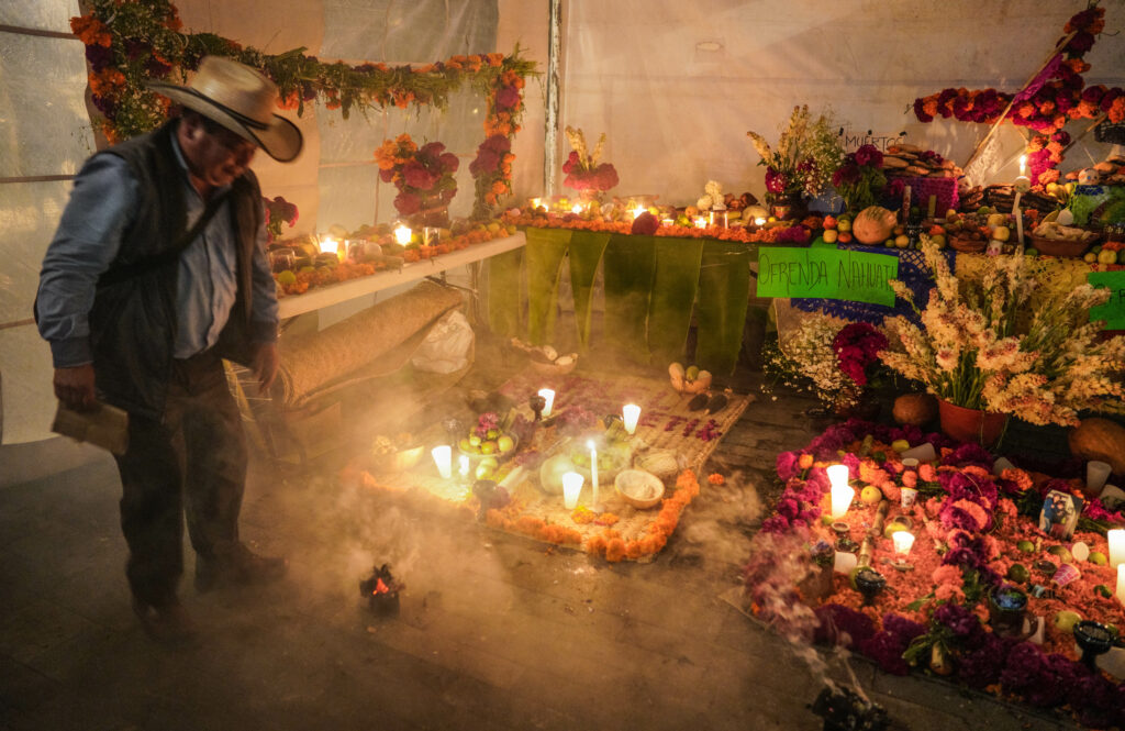 A person in a cowboy hat, vest, and boots stands next to flower clusters and garlands, burning candles, and a green sign that reads “Ofrenda Nahuatl.”
