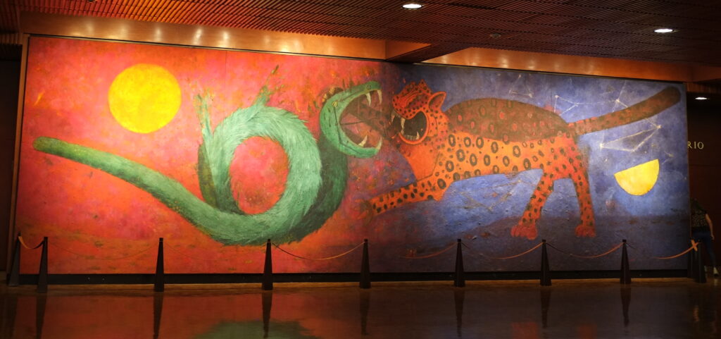 A large mural depicts a snake and jaguar confronting one another, their teeth bared. The snake is set against a red background with a sun; the jaguar is against a blue background with a half moon and stars.