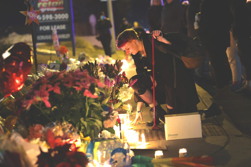 A photograph shows a crying person using a red cane to prop themselves up as they kneel in front of an assortment of flower bouquets, burning candles, and balloons arranged neatly on a sidewalk.