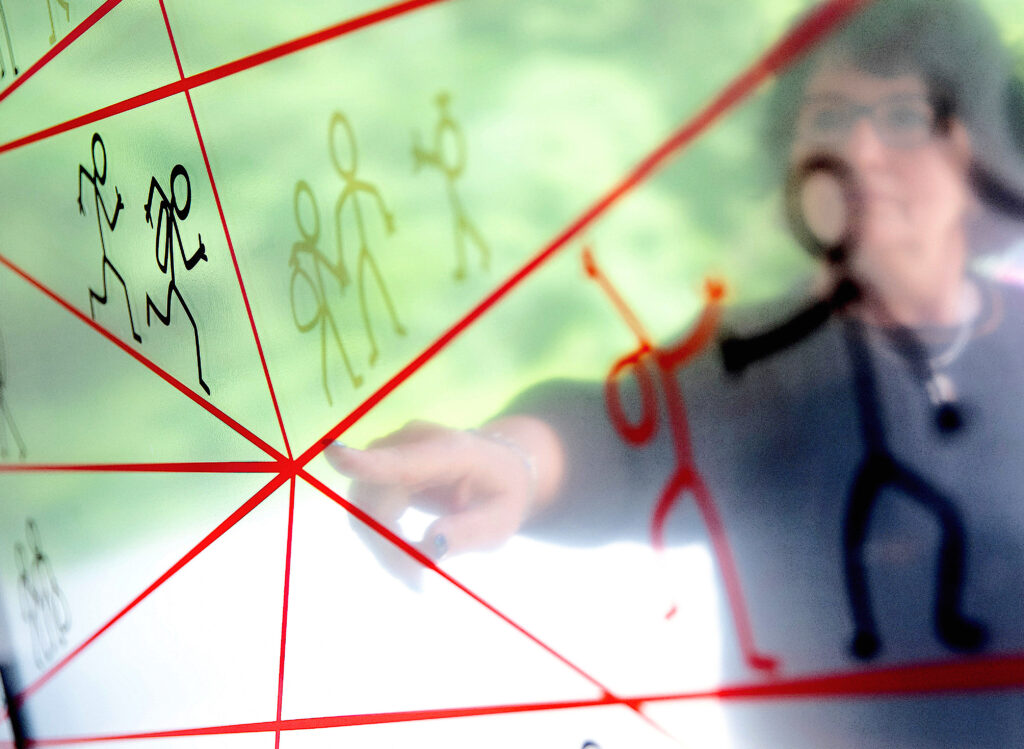 A photograph shows, through glass, a person with short black hair wearing glasses, a necklace, and a black shirt pointing at a graphic of stick figures. Different clusters of stick people interact in different scenarios as various red lines surround them, with several lines intersecting at a central node.