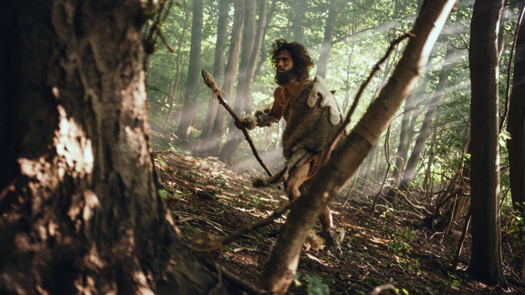 A photograph features a man walking in a shadowy forest with sun shining in from the right side. He has disheveled brown hair and a beard, wears a fur pelt, and holds a wooden spear with a stone tip.