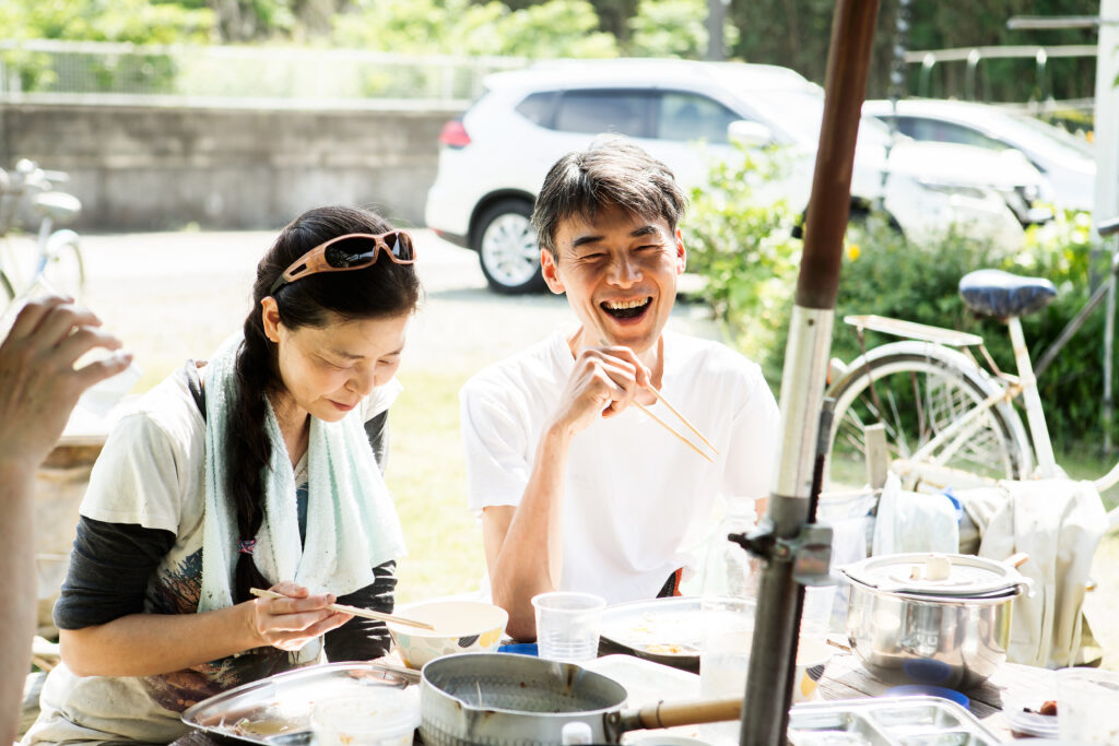 A photograph features two people smiling and holding chopsticks. Surrounded by sunlight, they sit side-by-side at an outdoor table with plates, pots, and plastic cups on it.