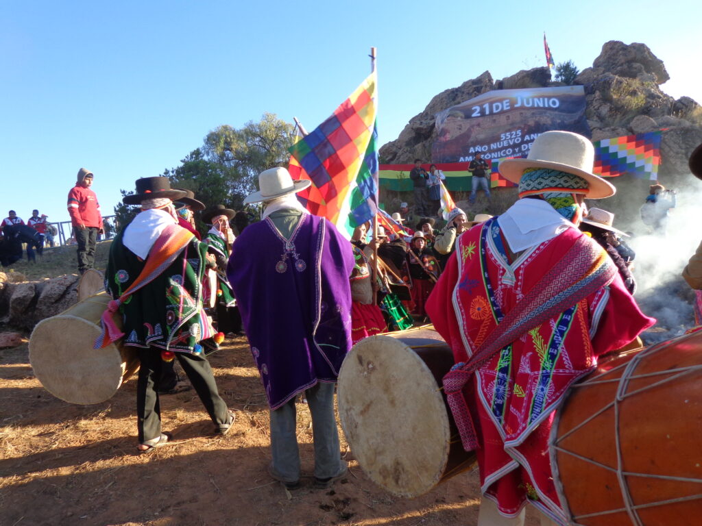 A photograph features a group of people in colorful ponchos and brimmed straw hats waving colorful flags and holding large drums. A banner in the distance reads “21 de Junio."