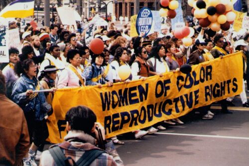 A photograph features a large crowd of people marching in a street. People in the front row hold up a large, long yellow banner with black text that reads “WOMEN OF COLOR FOR REPRODUCTIVE RIGHTS.” Other people also carry signs with different slogans.