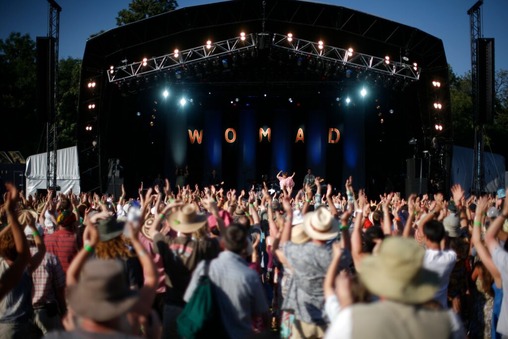 A photograph features a large crowd at a concert waving their hands in front of a dark stage lit by directional overhead spotlights and illuminated letters that spell out “WOMAD.”