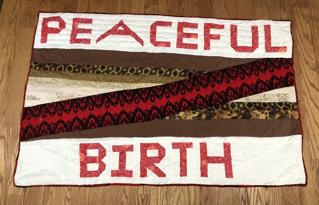 A quilt is spread out on a wooden floor. Red embroidered text reads “PEACEFUL BIRTH,” and the two words are separated by strips of red, brown, and black fabric.