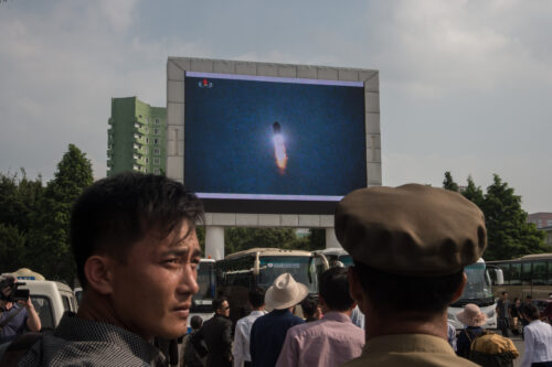 A crowd faces a large outdoor screen showing video of a nuclear missile launching into a blue sky. Several buses are visible in the background.