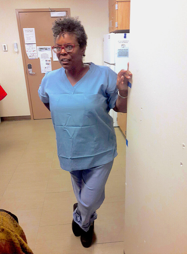 A woman wearing glasses and blue scrubs leans against a wall with her left hand while crossing one ankle over the other. She looks off camera right.