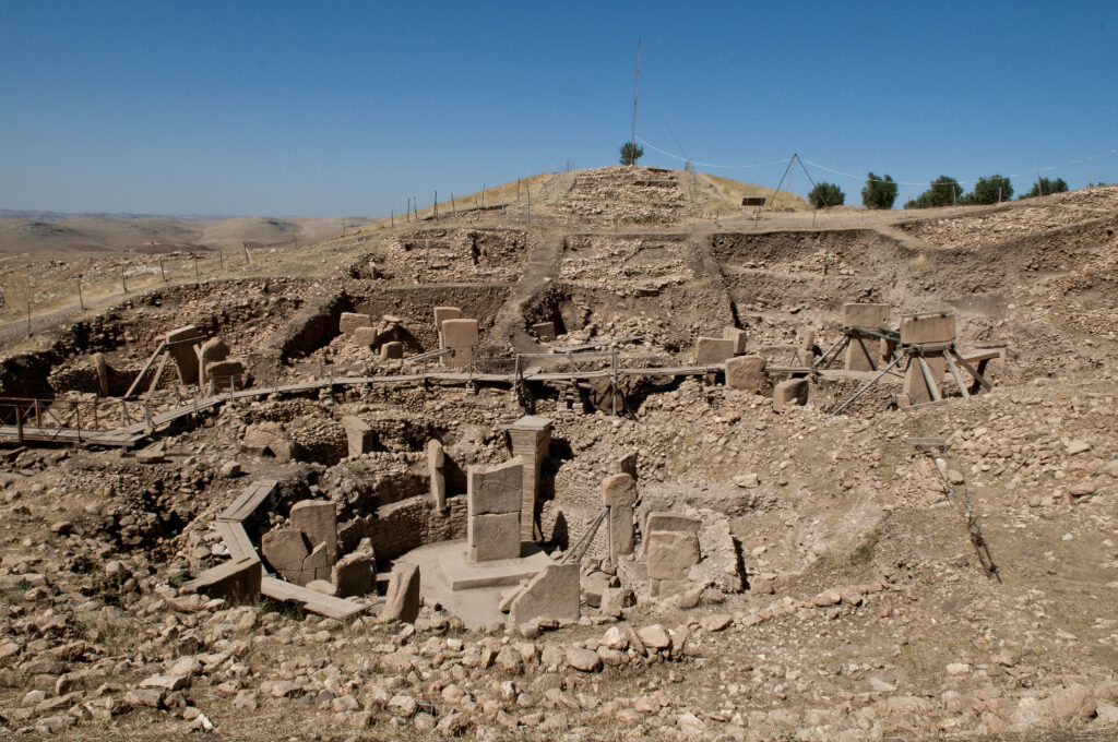 A photograph features ruins of brown stone and dirt in a circular arrangement. Behind this, the terrain ascends to a hill topped with green trees and blue skies.