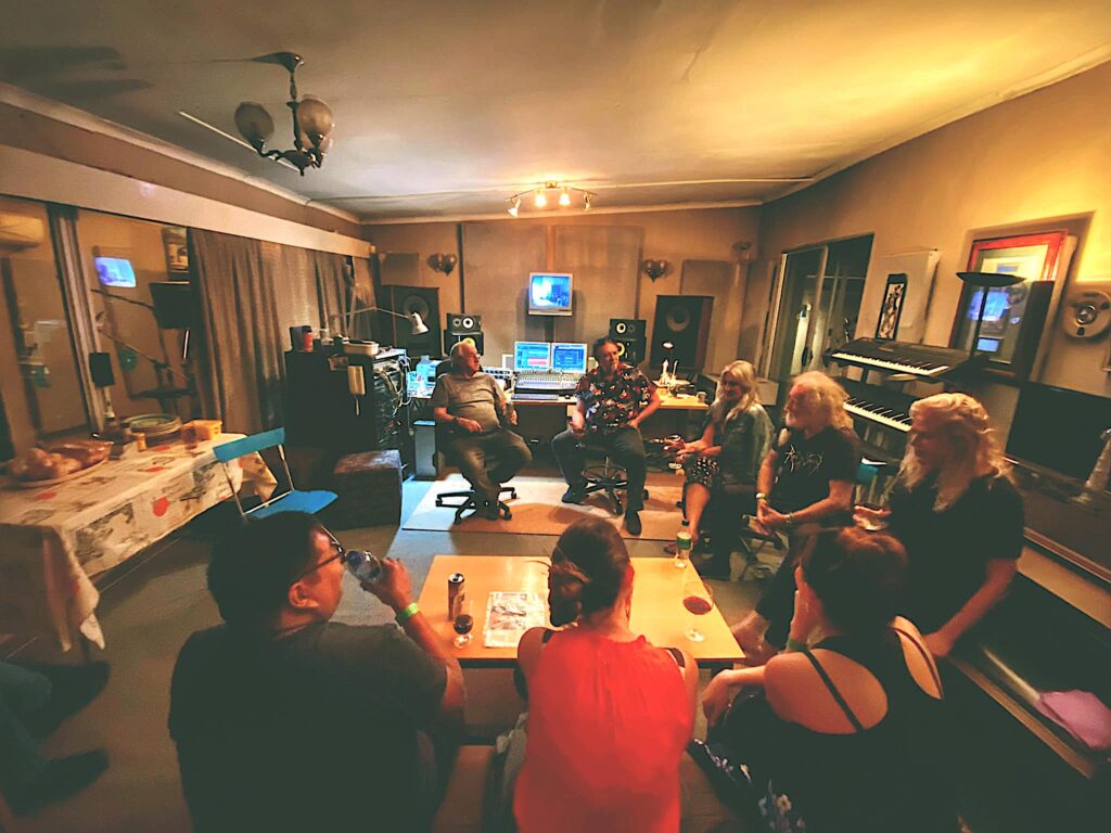 A photograph features several people in a room seated in a circle facing and conversing with one another. Electronic piano keyboards and open laptops line the room’s walls.