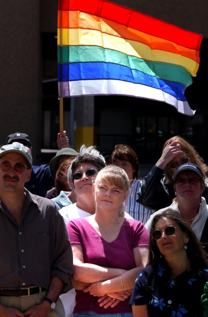 A photograph features a crowd of people looking straight ahead. A person with straight blonde hair wearing a burgundy T-shirt crosses their arms as someone behind them waves a rainbow flag overhead.