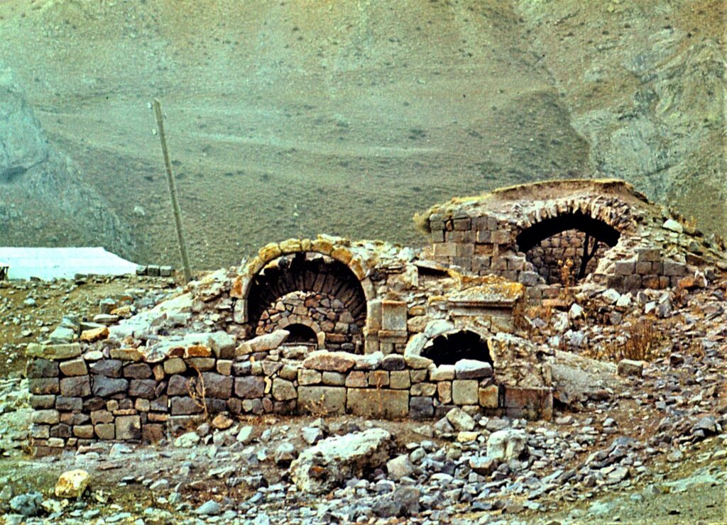 Two partially ruined domed structures built from beige and gray stones are surrounded by a wall made of the same materials.